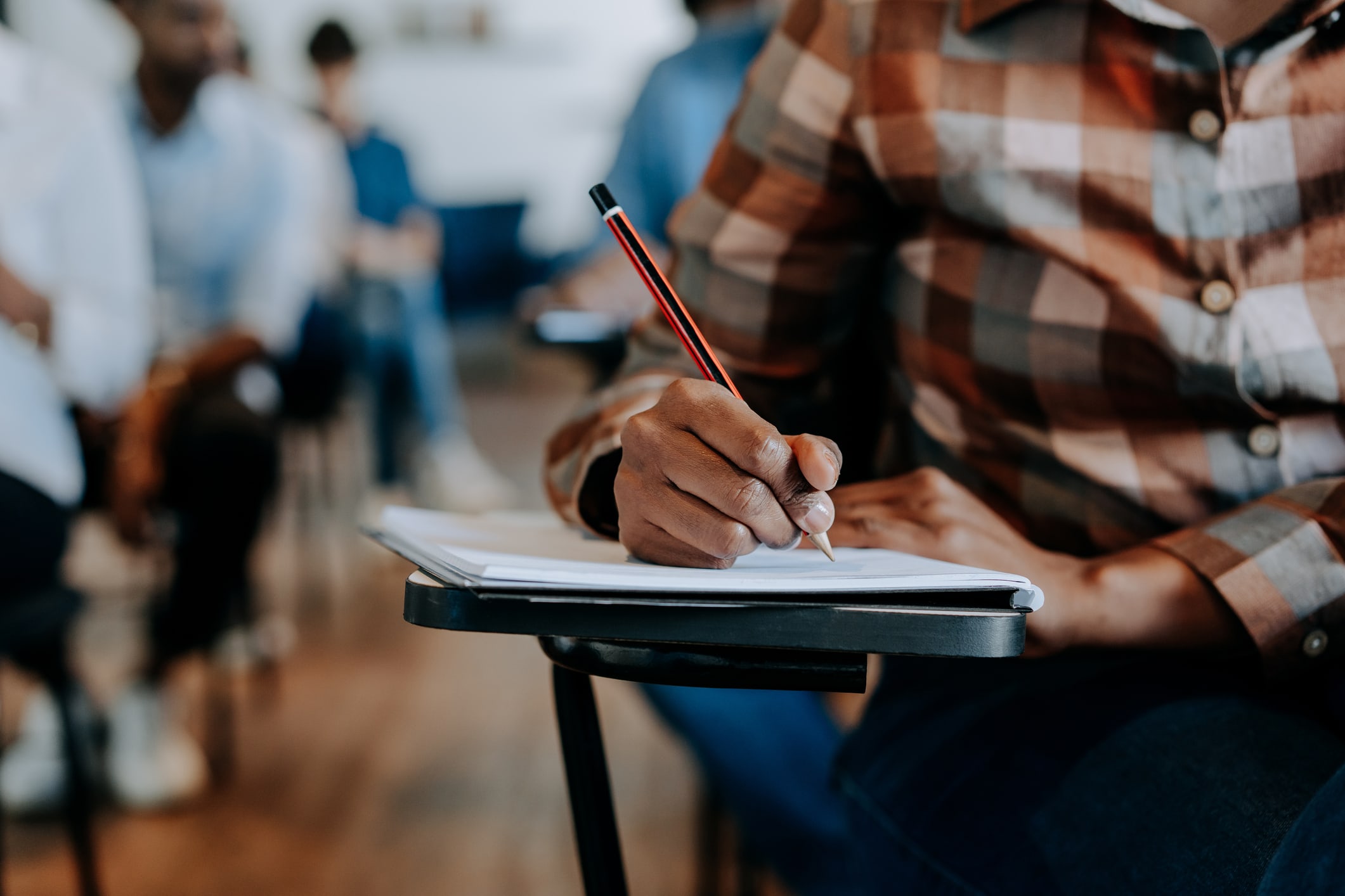 Close up of a student’s hand, taking notes with pen and paper in a classroom setting. A blurred background shows other students sitting at desks nearby.