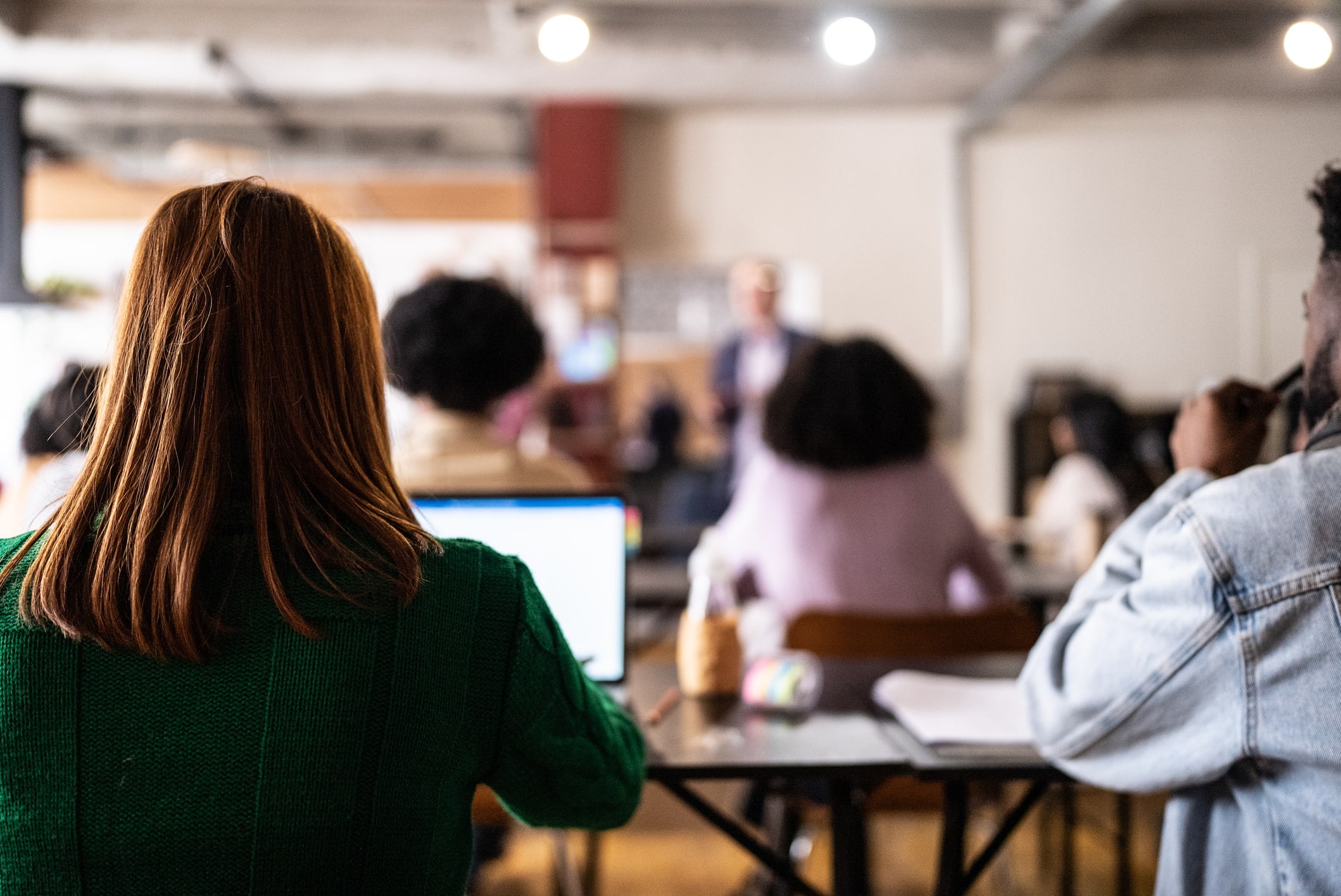 A view of a crowded classroom from the last row of seats. The background is blurred; the foreground shows a woman with her back to the camera, taking notes on her laptop.