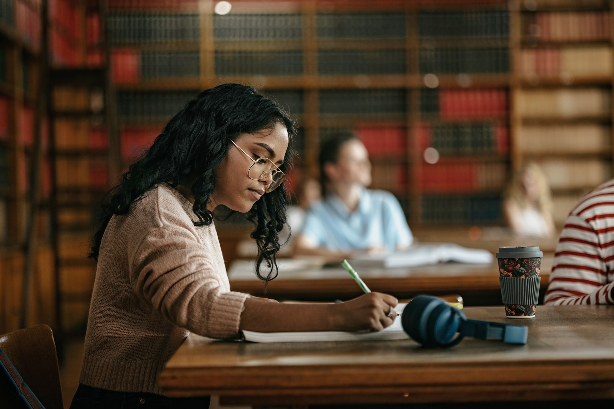 A woman is seated at a table in a library, writing with a pen and paper. Her coffee and headphones are nearby. 