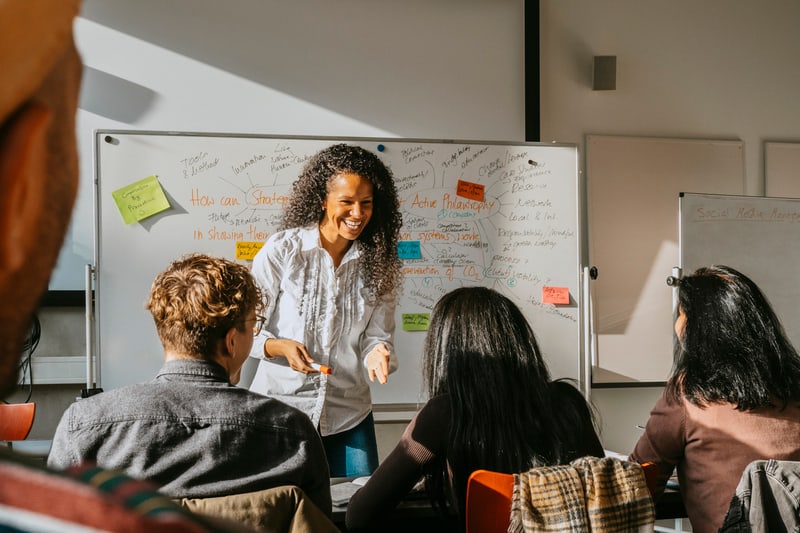 A female teacher is standing in front of a whiteboard, holding a marker and smiling at her students