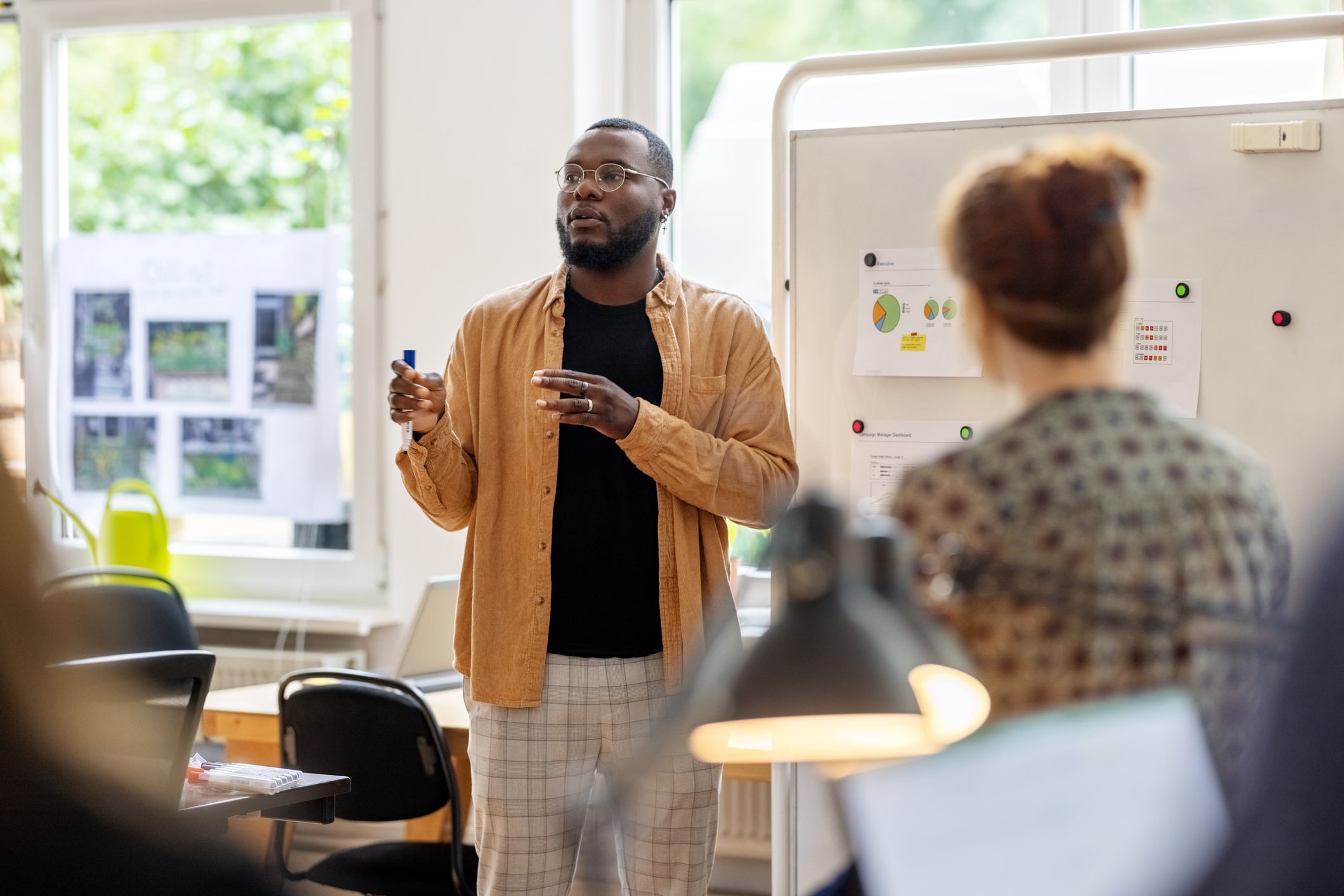 A young Black man gives a presentation to his colleagues in front of a white board with charts and graphs. He gestures with a dry erase marker as he speaks. 