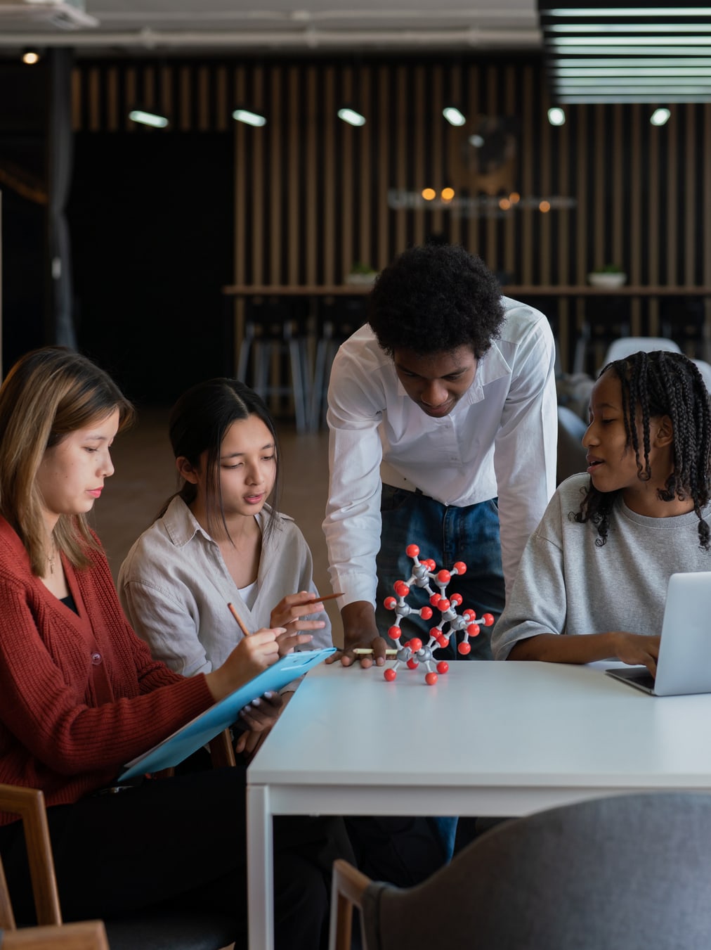 A group of researchers in a range of ages, genders, and races closely examine a 3D molecular model on a long table. One takes notes on a clipboard, another is using her laptop.
