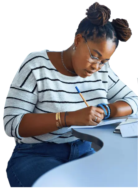 A young woman in a striped shirt is sitting at a table,  concentrating closely and writing on a worksheet with a pencil. 